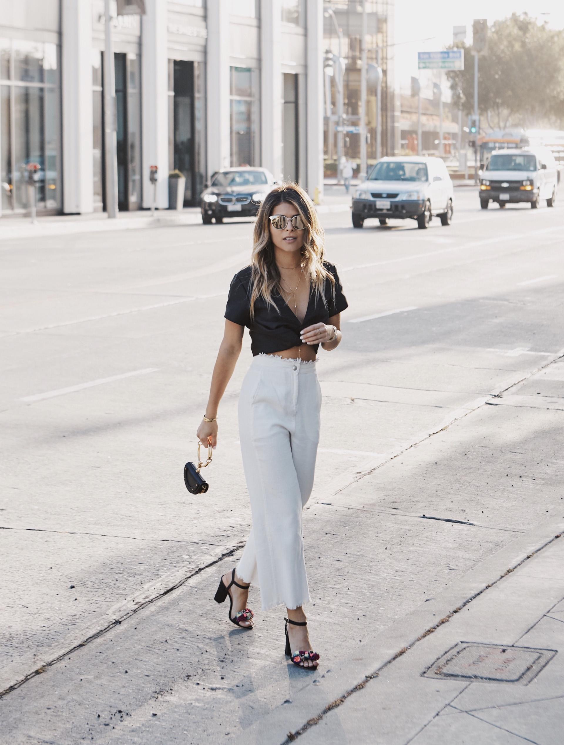 Wearing a black bra, white pants and sandals, standing by trees, a