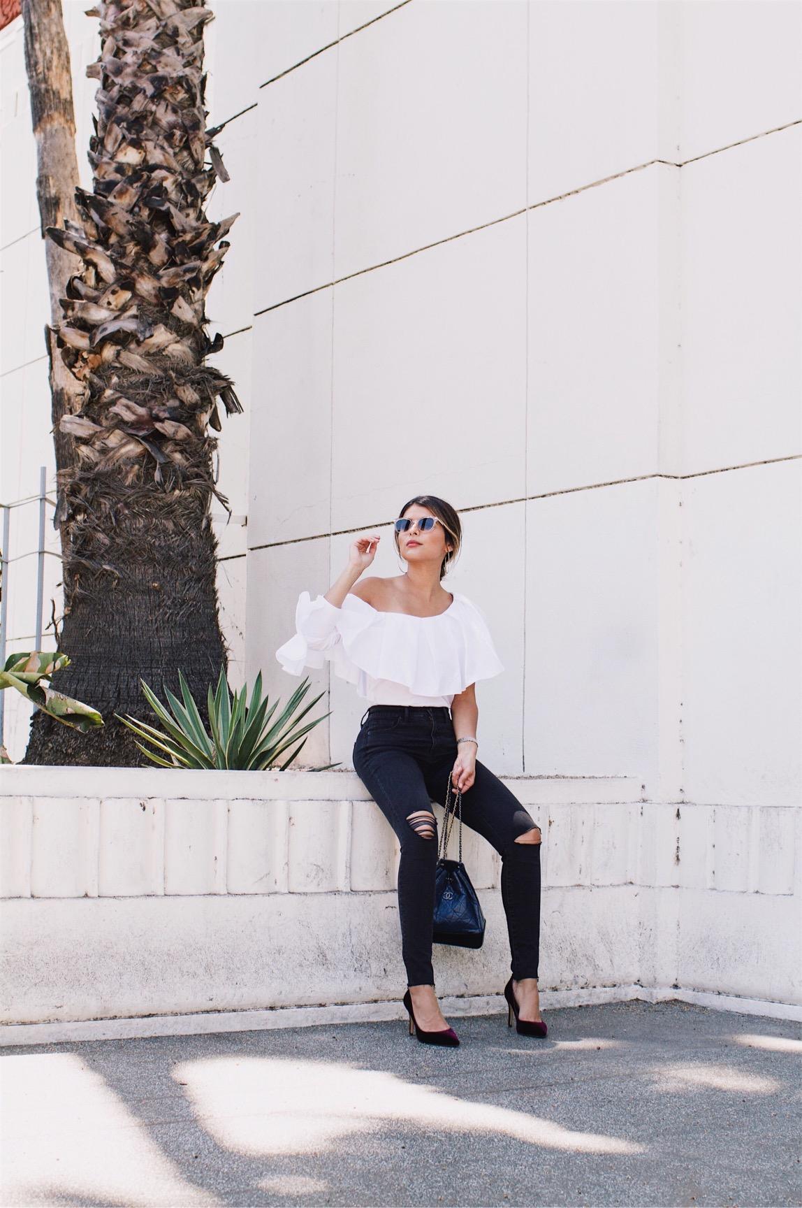 Pam Hetlinger wearing a White Ruffle Top styled with black ripped jeans and velvet pumps.