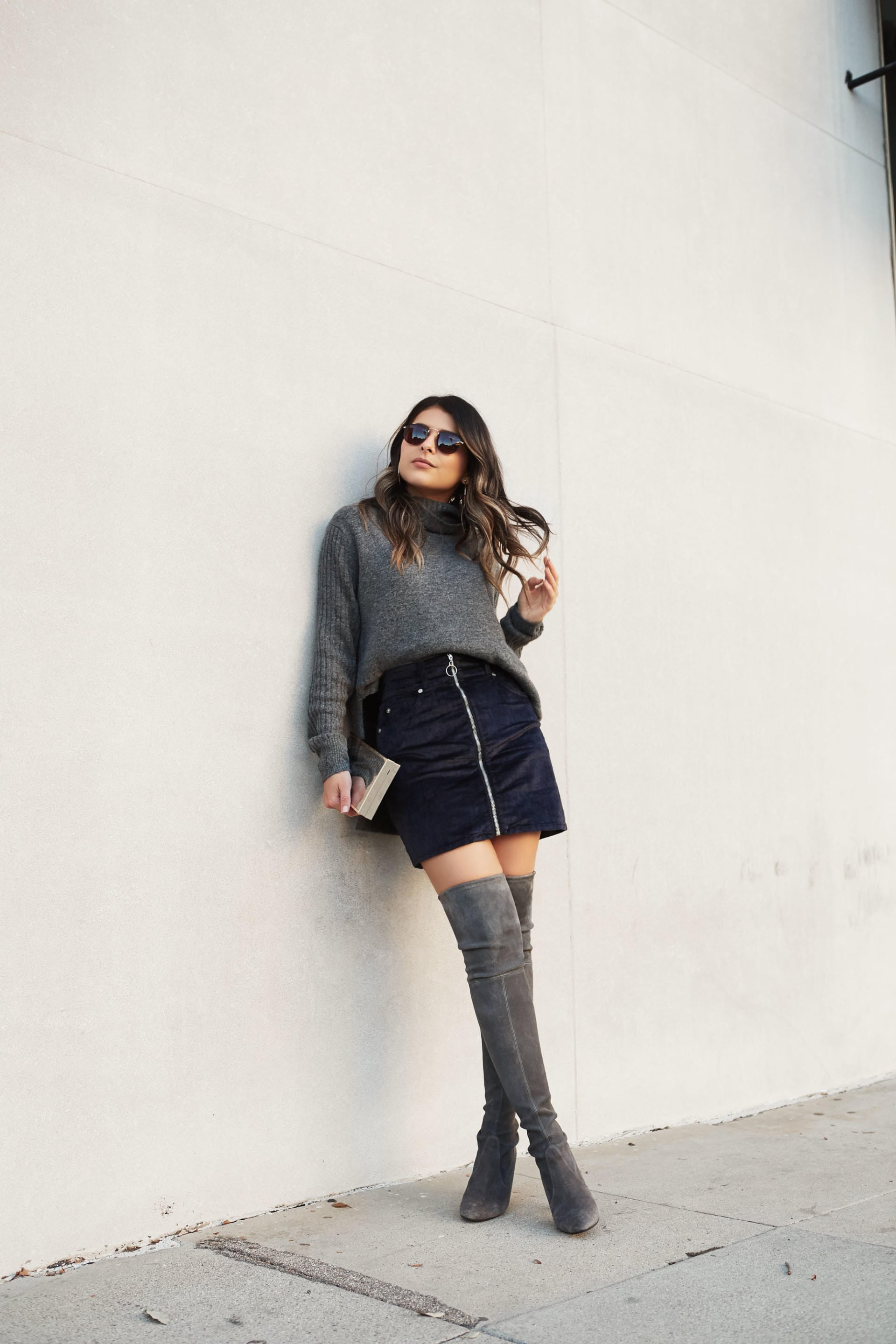 Pam Hetlinger of the fashion, beauty, travel, and lifestyle blog The Girl From Panama, shows how to transition a skirt into a holiday party ensemble by pairing it with thigh high boots and a gray sweater.